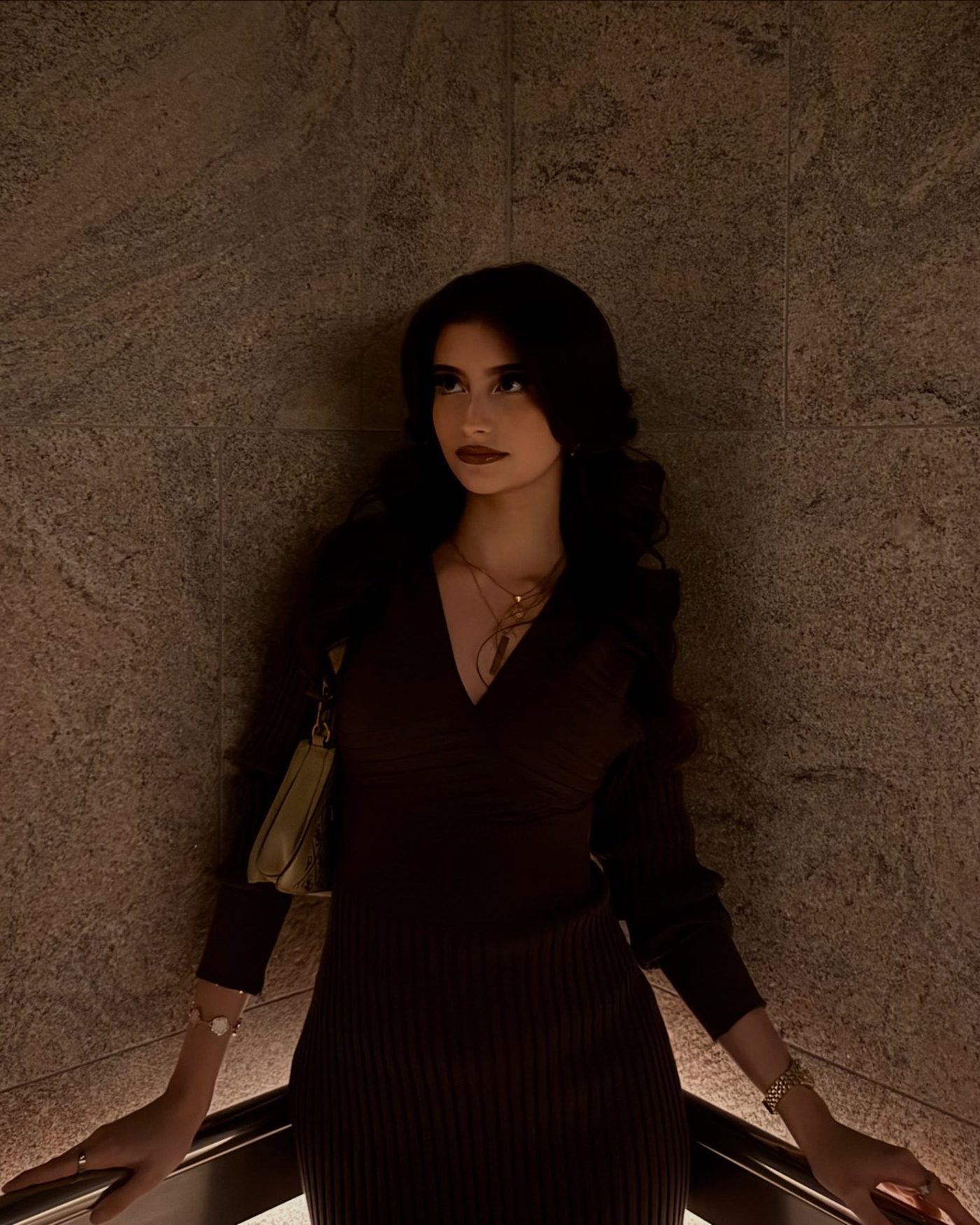 Aisha standing against a lit stone wall in a fitted espresso knit dress, looking past the camera.